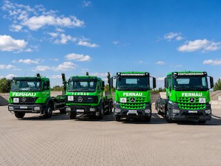 Vier grüne Fernau-Containerdienst-Lkw auf einem gepflasterten Betriebshof unter blauem Himmel. @ 