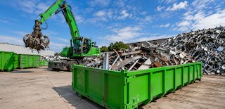 Grüner Abrollcontainer mit Altmetall auf einem Recyclinghof, während ein Umschlagbagger Metallschrott sortiert. @ 