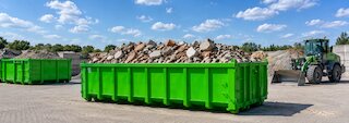Grüner Abrollcontainer mit Bauschutt auf einem Recyclinghof, daneben ein Radlader und weitere Container. @ 