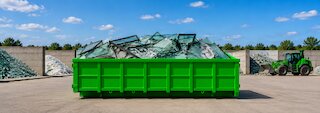 Grüner Abrollcontainer mit sortierten Glasabfällen auf einem Recyclinghof, im Hintergrund getrennte Glasbestände und ein Umschlagbagger. @ 