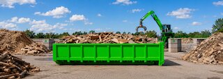Grüner Abrollcontainer mit sortierten Holzabfällen auf einem Recyclinghof, im Hintergrund arbeitet ein Umschlagbagger. @ 