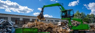 Grüner Umschlagbagger sortiert Holzabfälle auf einem Recyclinghof, daneben befinden sich getrennte Metallreste. @ 