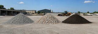 Drei getrennte Materialhaufen aus grauem Betonrecycling, rötlichem Bauschuttmineral und dunklem Mutterboden auf einem Recyclingbaustoffhof. @ 