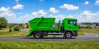 Grüner Container-Lkw mit Absetzcontainer auf einer regionalen Straße vor Feldern, Gewerbegebieten und einem niedersächsischen Ortsrand. @ 
